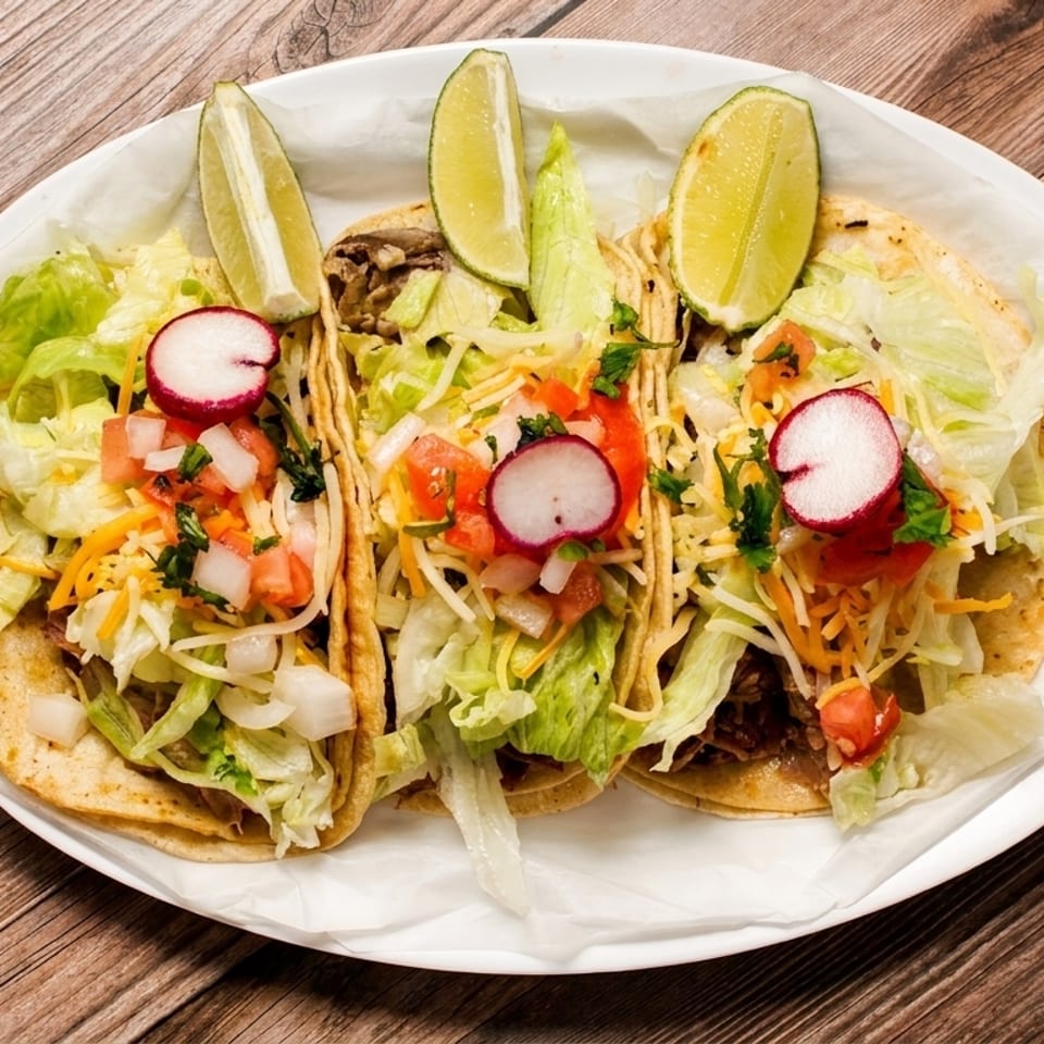 Three tacos with meat, lettuce, cheese, pico de gallo, radishes, and lime wedges on a white plate.
