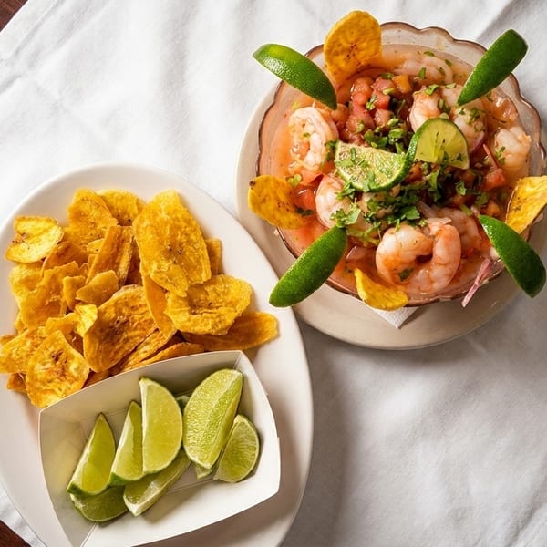 Shrimp ceviche with plantain chips and lime wedges on a white napkin over a wooden table.