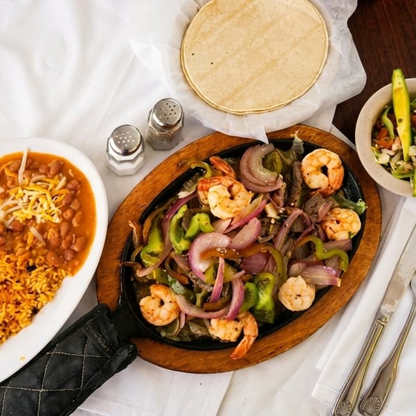 Shrimp fajitas with bell peppers and onions, rice, beans, tortillas, and avocado salad on a table.