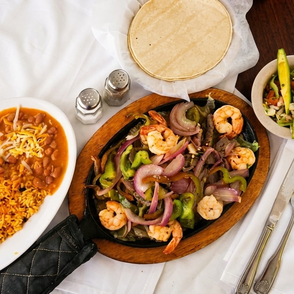 Shrimp fajitas with bell peppers and onions, rice, beans, tortillas, and avocado salad on a table.