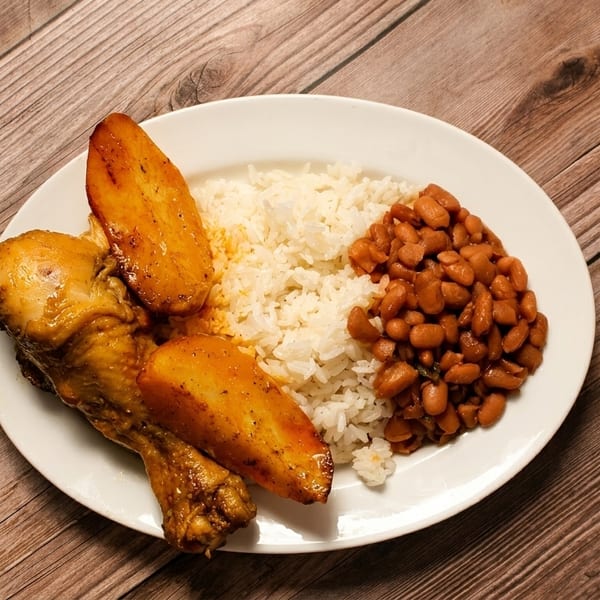 A white plate with roasted chicken, plantains, white rice, and beans on a wooden table.