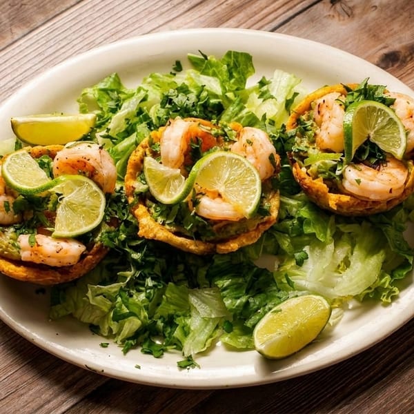 Three shrimp and avocado tostadas with lime wedges on a bed of lettuce, on a white plate.